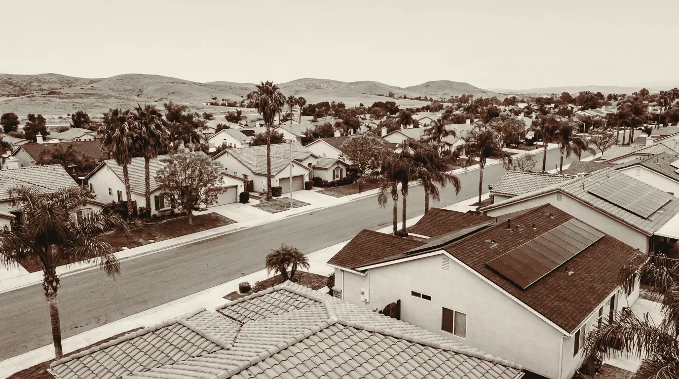 An elevated view over a Southern California residential street of single storey houses with tile and shingle roofs, palms and jacarandas along the kerb, rooftop solar on several of the houses, and dry hills on the horizon.