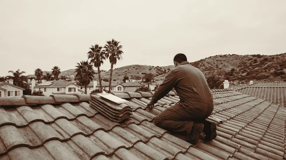 A worker in a hard hat kneels on a clay tile roof taking a measurement with a tape, a clipboard beside him, with suburban rooftops, palms and dry hills behind.